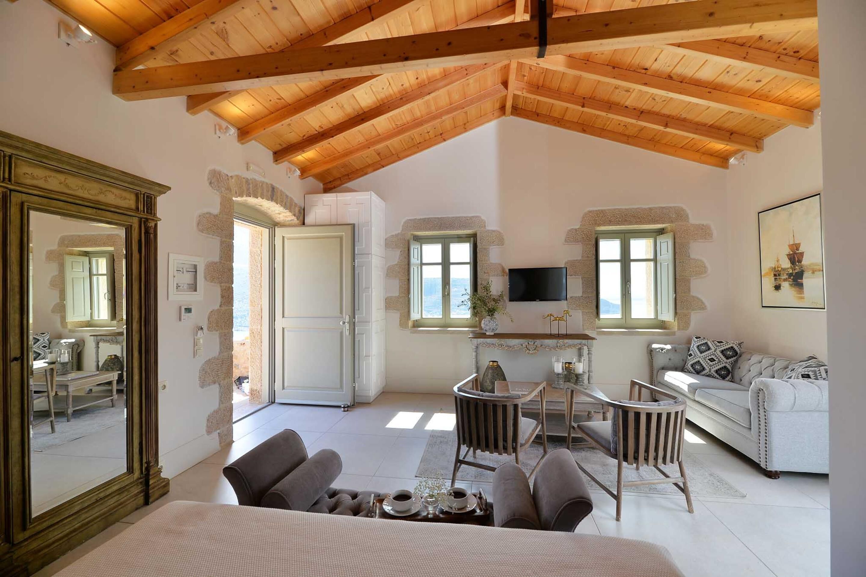 A spacious room with a high wooden ceiling, a large mirror, a dining area with a table and chairs, a sofa, and a TV mounted on the wall.