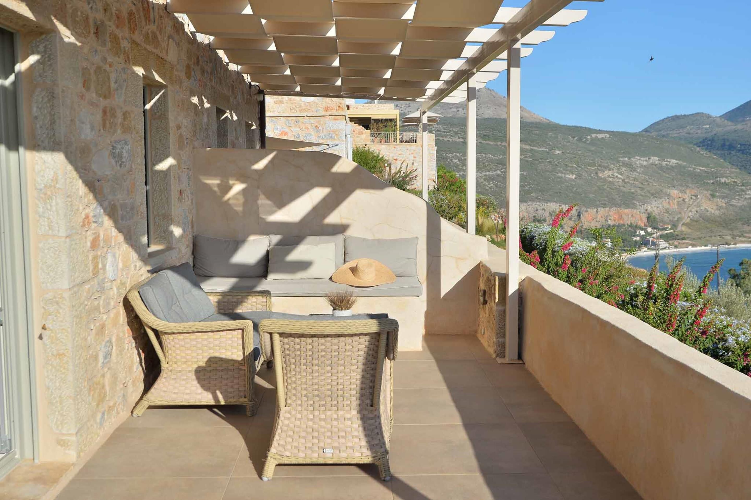 A balcony with wicker furniture, a stone wall, and a view of the sea and hills.