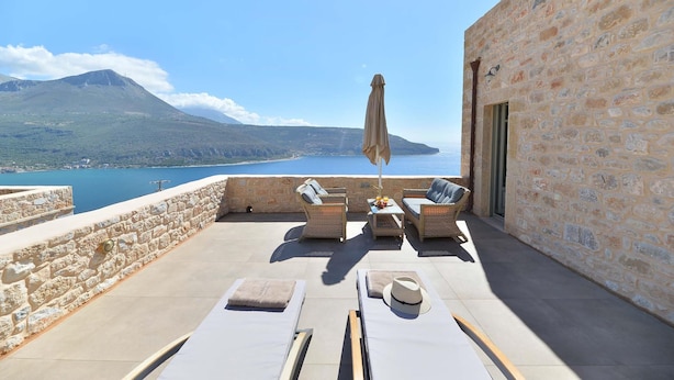 Una terraza con muros de piedra, sillones de descanso, una sombrilla y vistas a la montaña y al mar.