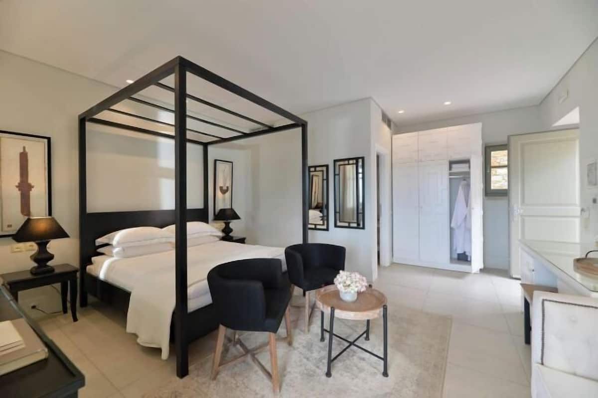 Un dormitorio moderno con cama con dosel, dos sillas negras, una mesita redonda y un baño blanco visible a través de una puerta abierta.