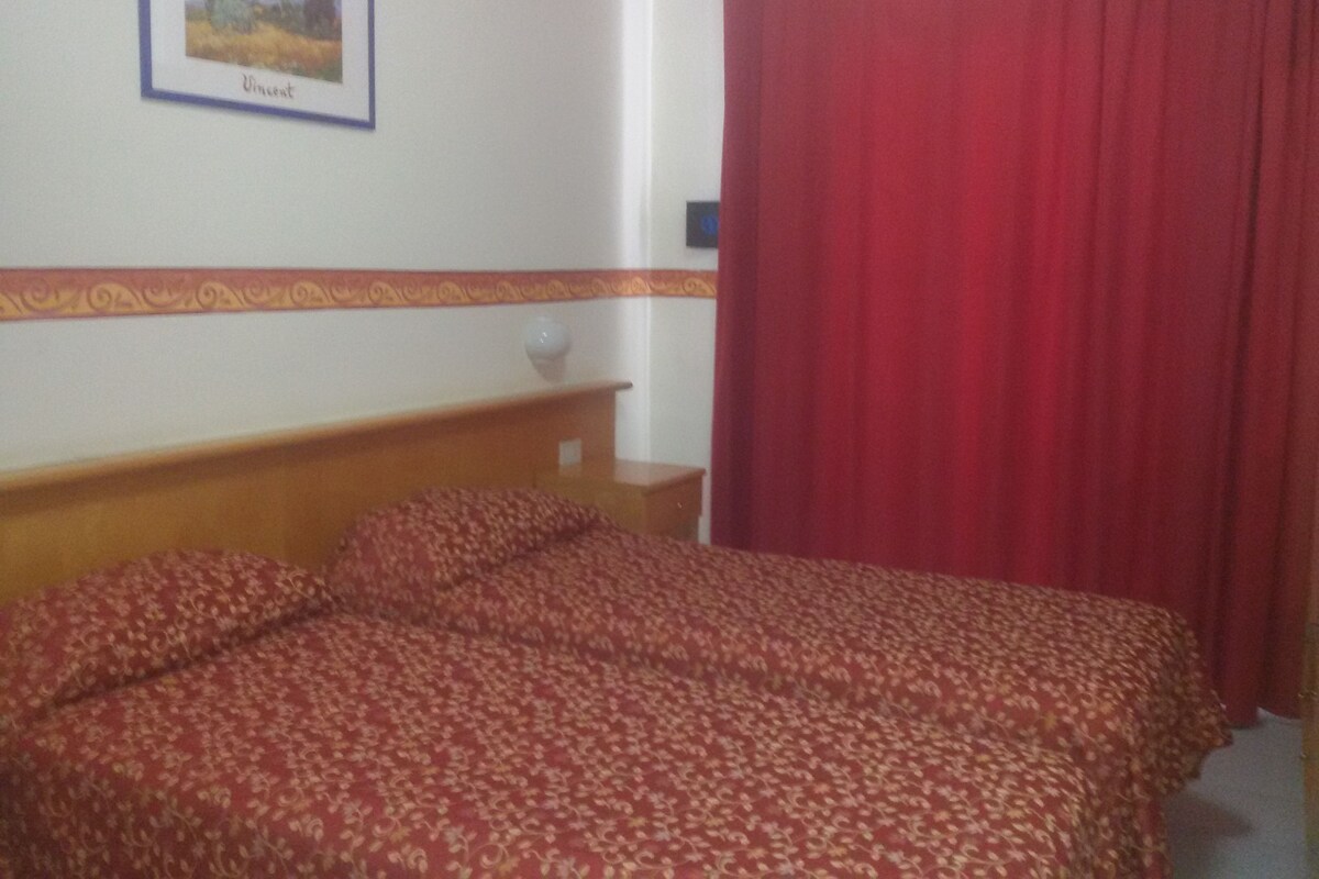 A bedroom with a bed, a nightstand, a framed picture on the wall, and a red curtain.
