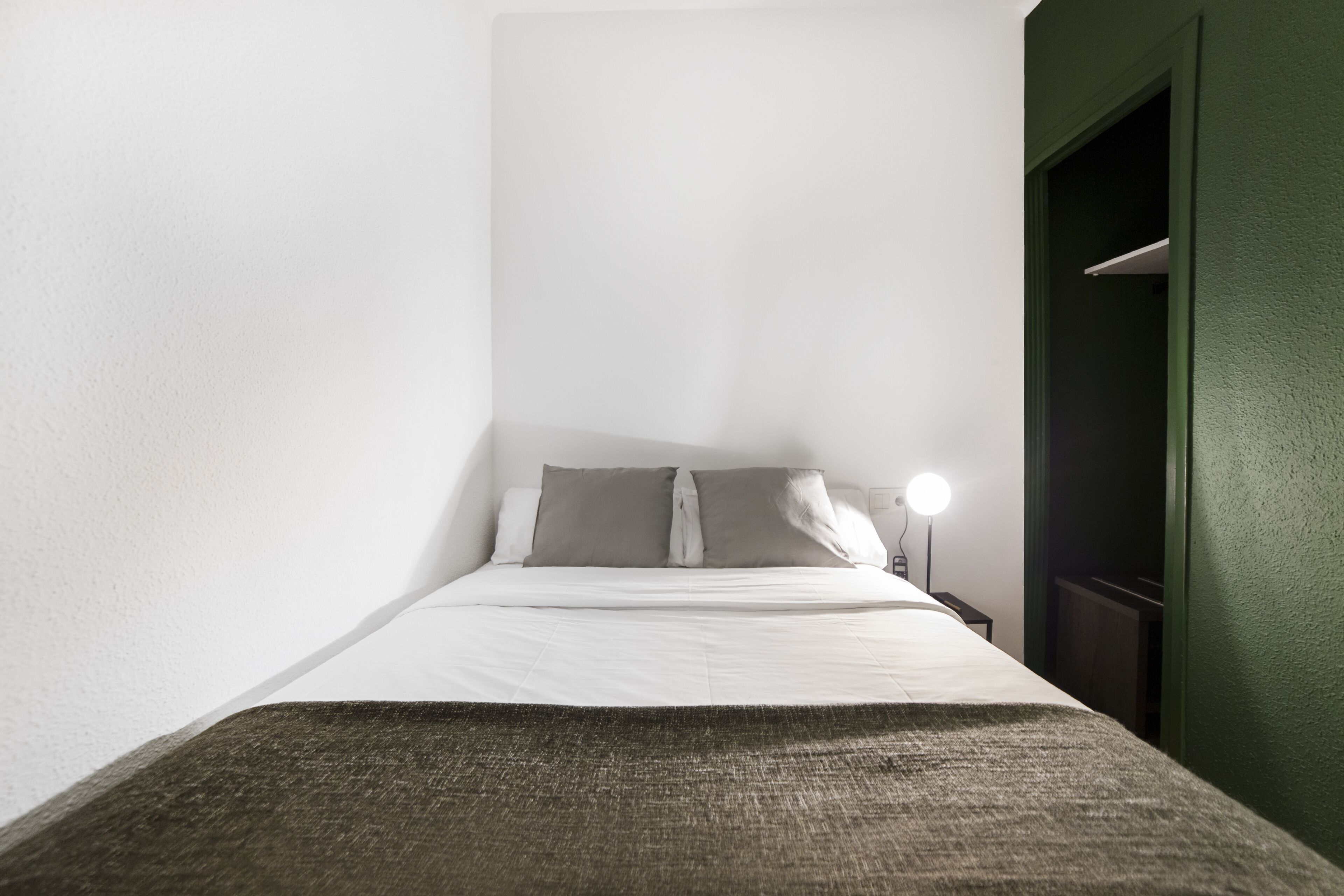 A small, neatly made bed with white linens and two pillows, a bedside lamp, and a green wall.