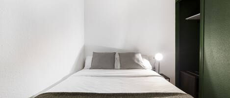 A small, neatly made bed with white linens and two pillows, a bedside lamp, and a green wall.