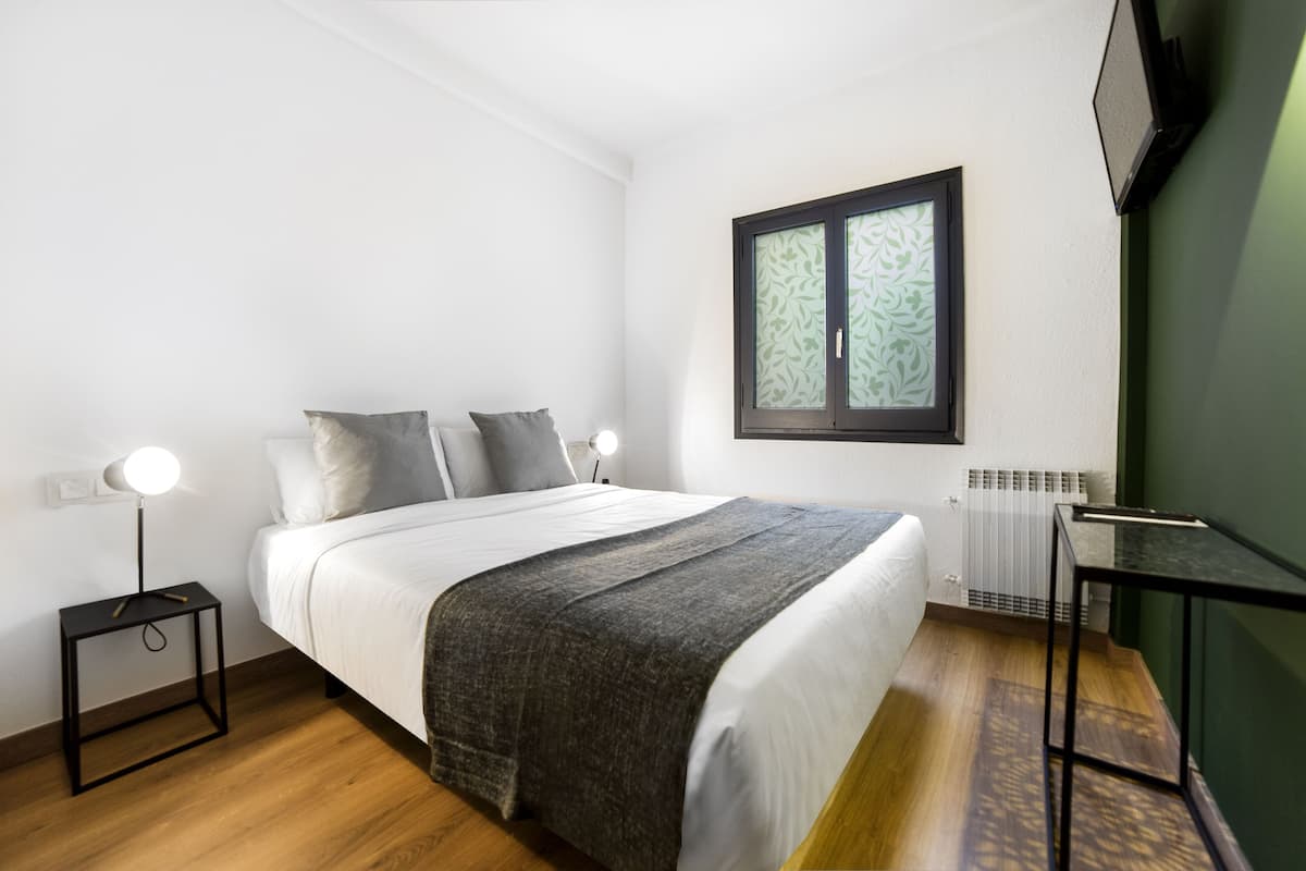 A bedroom with a bed, bedside table, lamp, radiator, and a window with a decorative frame.