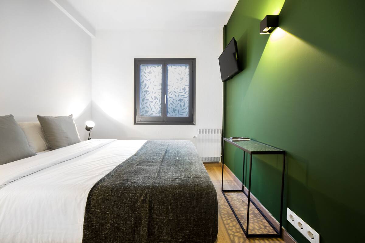 A bedroom with a bed, a nightstand, a wall-mounted TV, and a window with frosted glass.