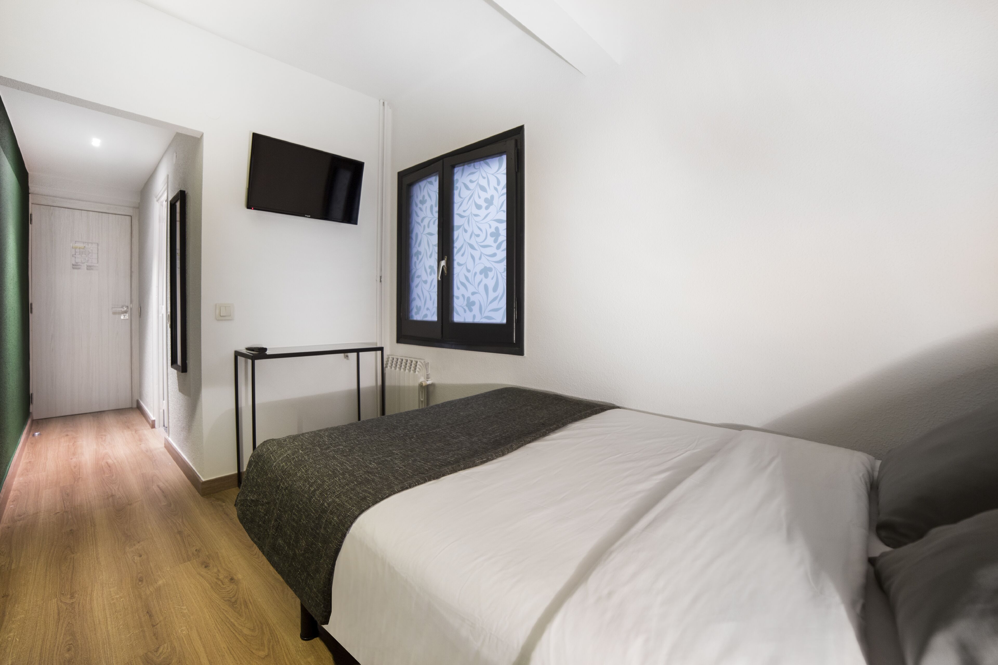 Un dormitorio con cama, televisor montado en la pared y ventana con vidrio esmerilado.