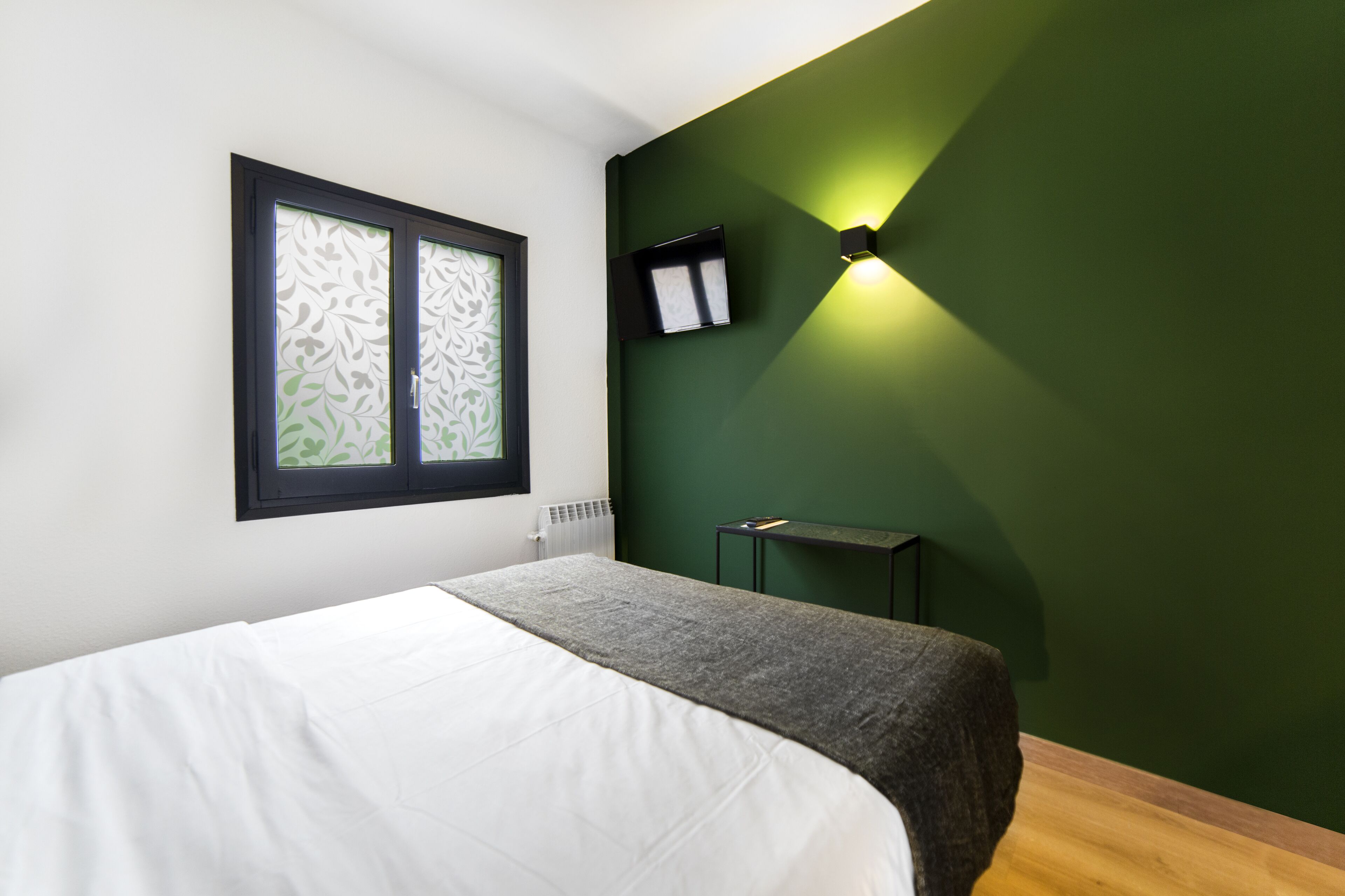 Une chambre à coucher avec un lit, une fenêtre ornée d’un motif décoratif, un luminaire fixé au mur et un radiateur.