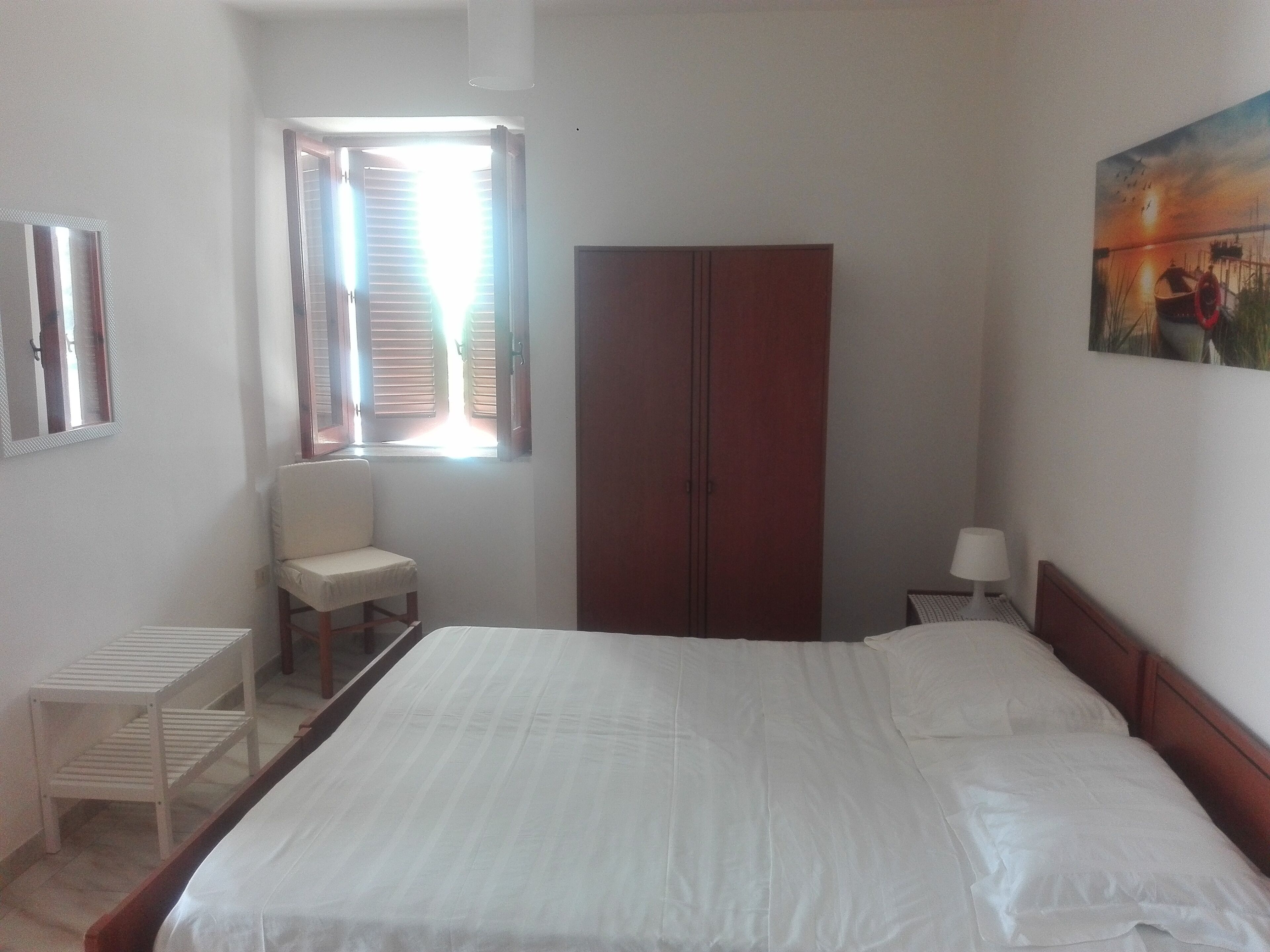 A bedroom with a bed, a chair, a wardrobe, a window with blinds, and a painting on the wall.