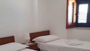 Een kamer met drie eenpersoonsbedden, een houten hoofdbord, een nachtkastje met een lamp en een raam met jaloezieën.