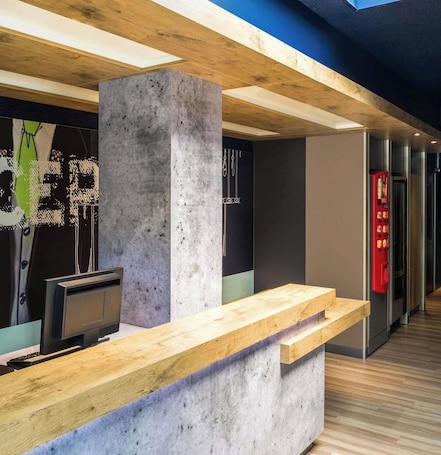 A reception desk with a wooden counter and a concrete pillar, a vending machine, and a modern elevator.. ibis budget Nantes Nord Saint-Herblain