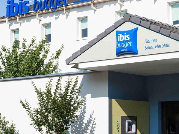 A building with 'ibis budget hotels' signage, a small ATM, and a tree in front. - ibis budget Nantes Nord Saint-Herblain (Saint-Herblain)