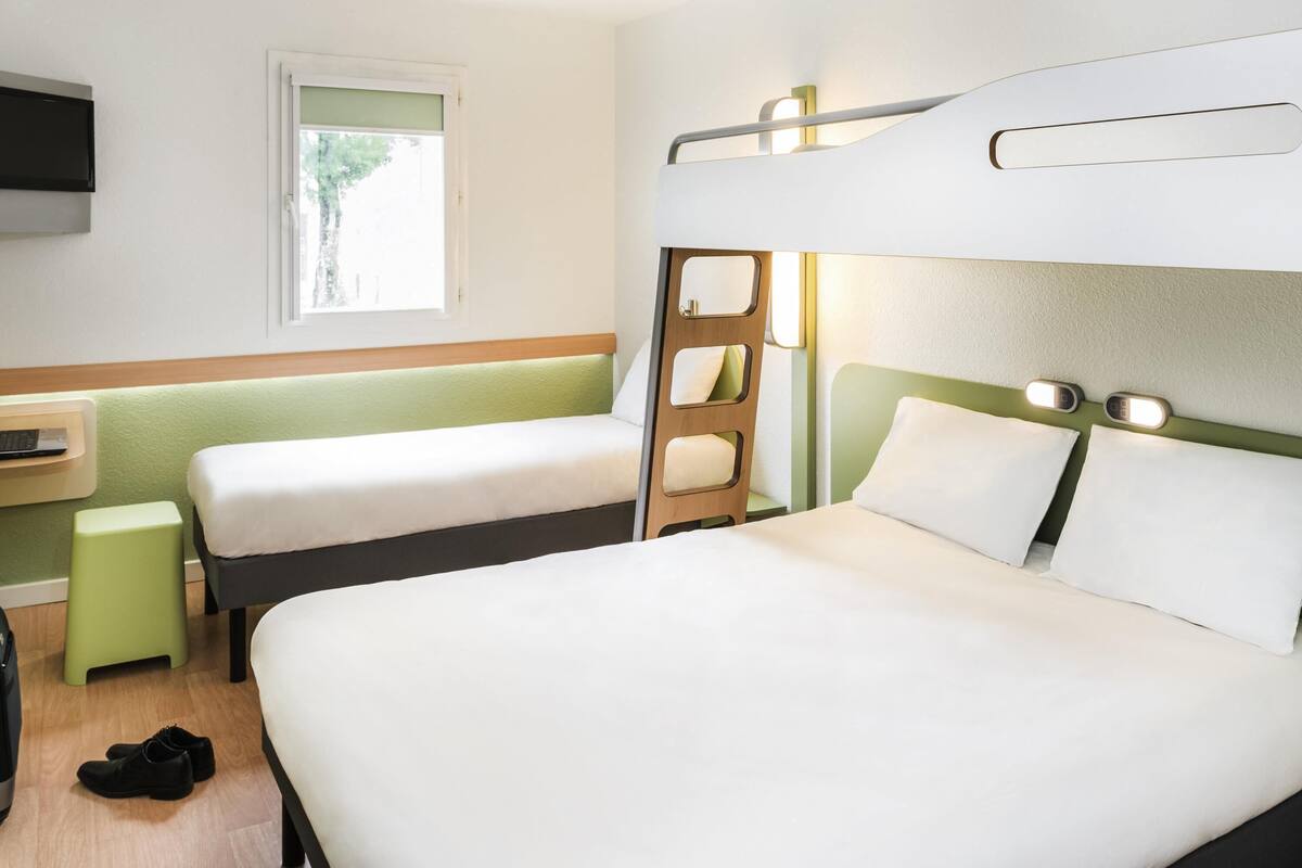 A hotel room with a bunk bed, a single bed, a desk with a chair, a TV mounted on the wall, and a window with a view of trees.