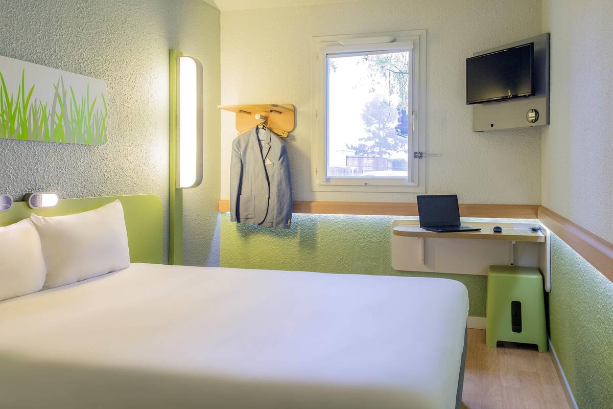 A hotel room with a bed, a desk with a laptop, a TV, and a window with a view of trees.