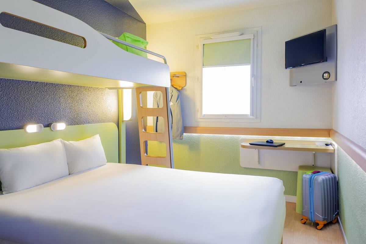 A hotel room with a bunk bed, a desk with a chair, a TV mounted on the wall, and a suitcase on the floor.