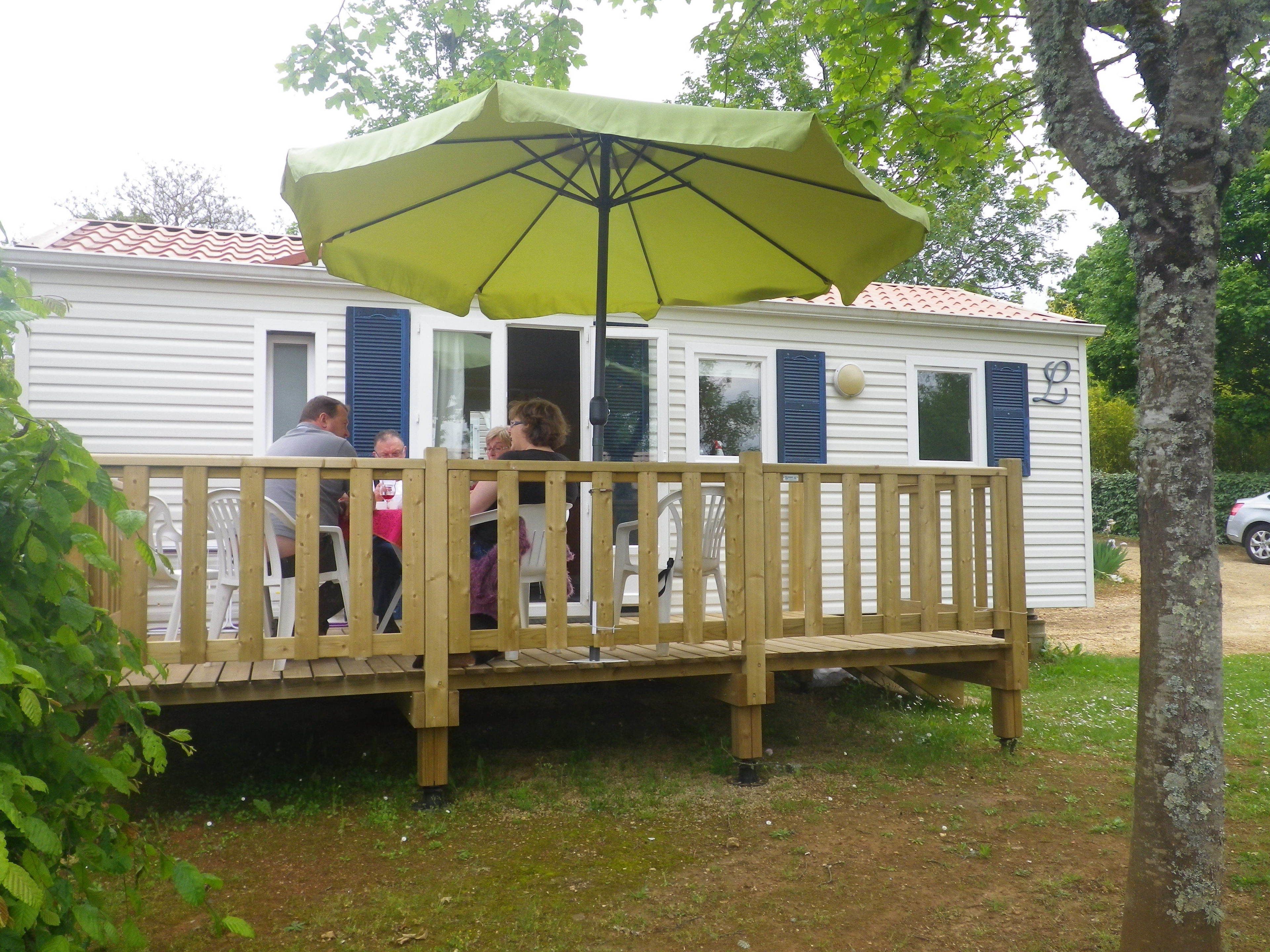 A wooden deck with a green umbrella, white walls, and blue shutters of a mobile home.