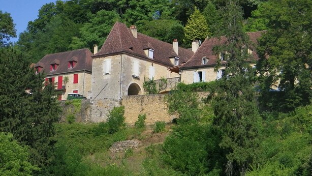 A historic building with multiple chimneys, a stone wall, and lush greenery.