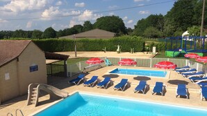 A swimming pool with blue water, surrounded by lounge chairs and umbrellas, and a small building in the background. - Camping Les Poutiroux - Tente Lodge (Limeuil)