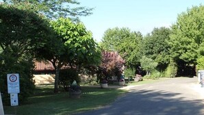 A paved driveway leading to a building with a red structure on the right. There are trees and a sign with text on the left. - Camping Les Poutiroux - Tente Lodge (Limeuil)