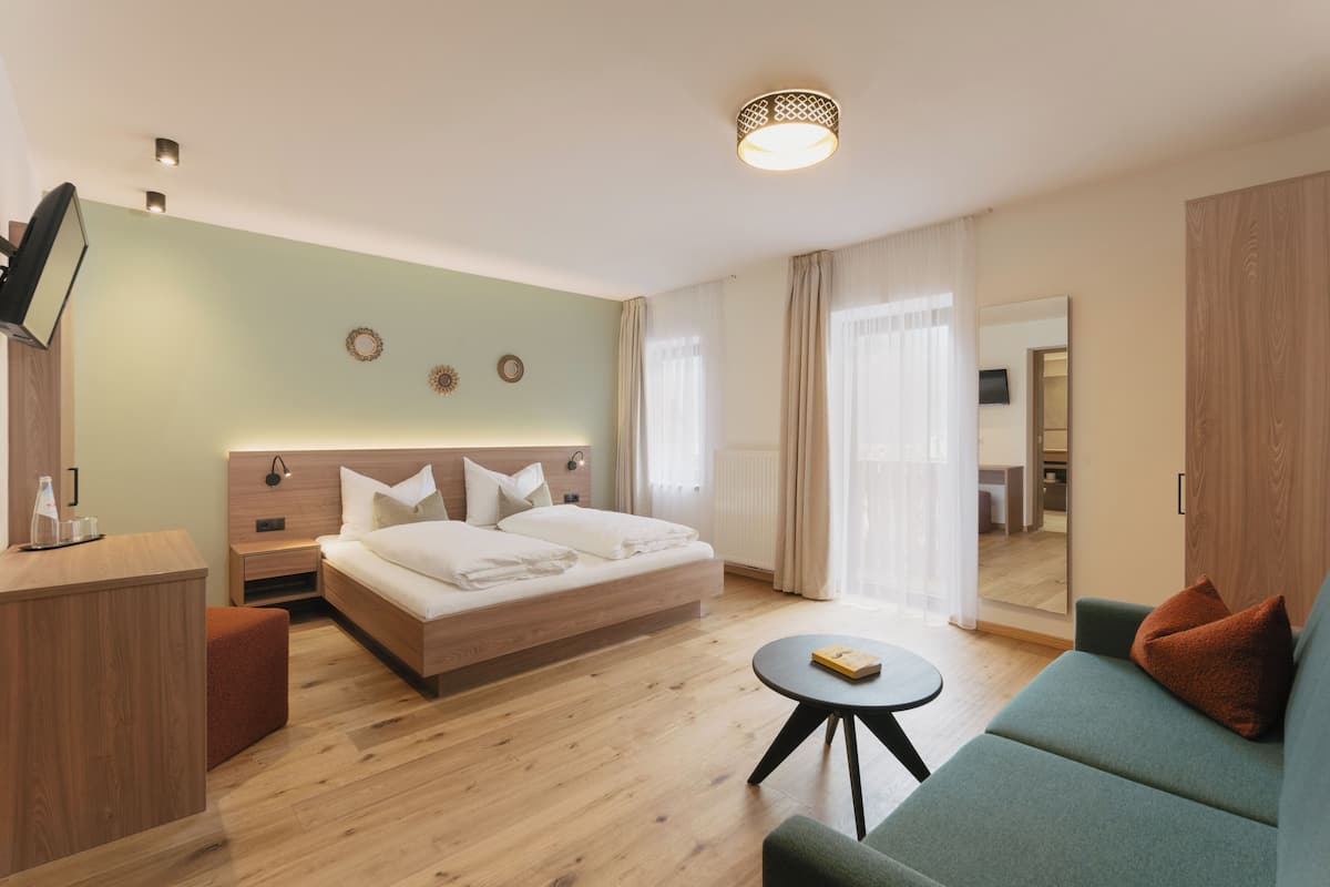 Habitación de hotel moderna con una cama de madera, un sofá verde, una mesita redonda y un televisor sobre un mueble de madera.