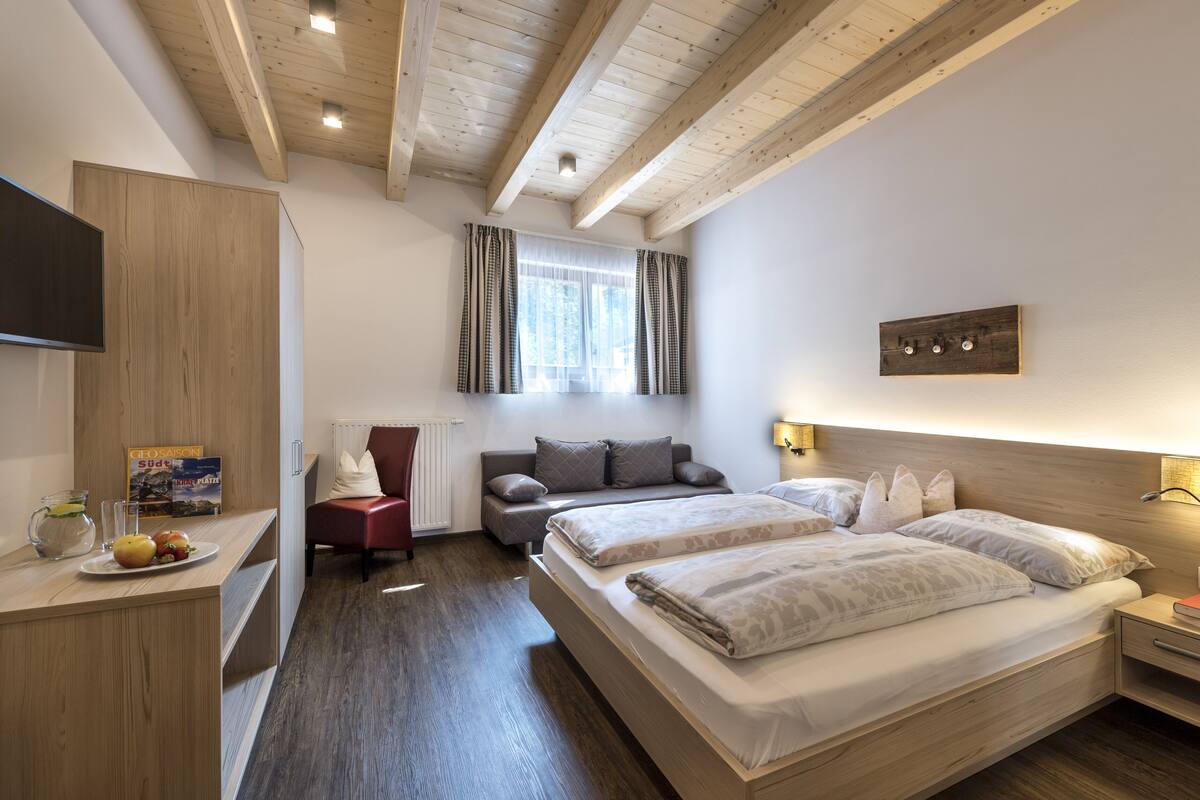 Un dormitorio moderno con techo de madera, una cama con dos almohadas, una mesita de noche con un libro y una ventana con cortinas.