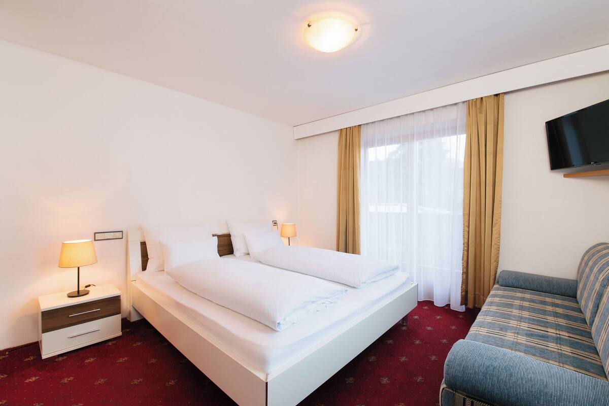 Habitación de hotel con cama, mesita de noche con lámpara, sofá, televisor montado en la pared y ventana con cortinas.