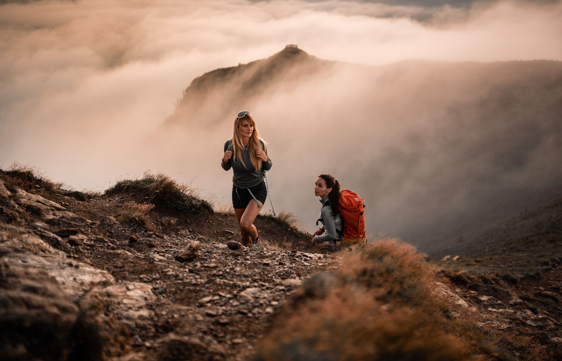 A mountainous landscape with two hikers, one standing and one kneeling, with a misty background.