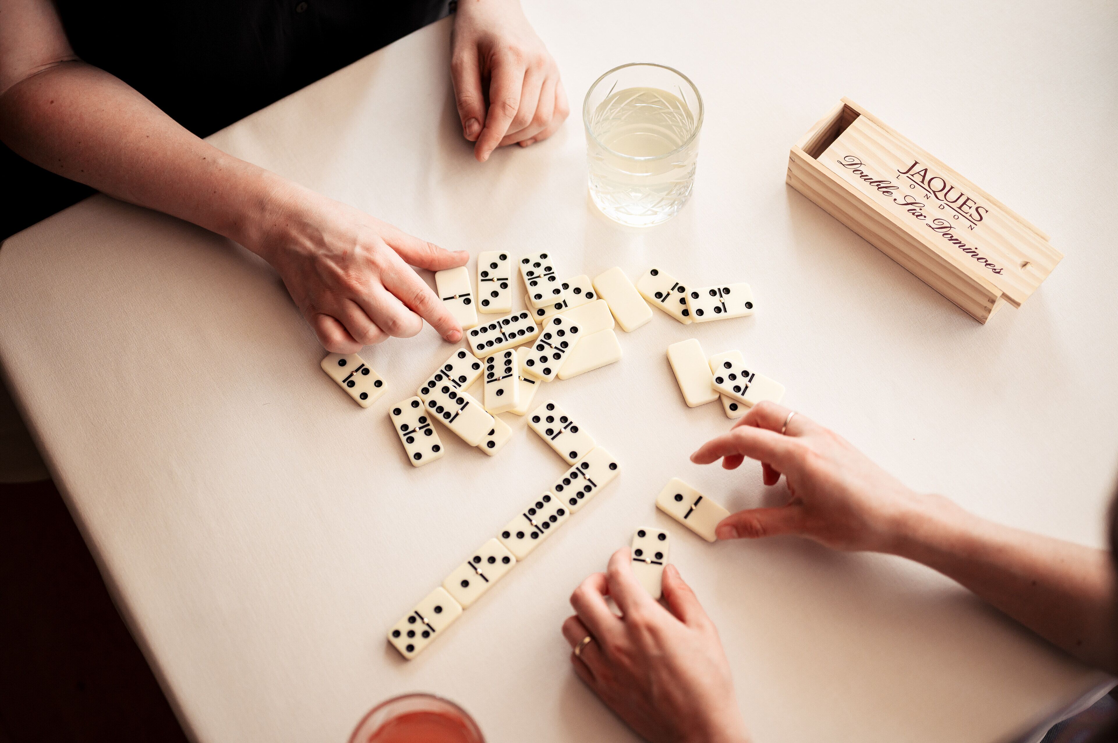 A table with dominoes, a glass of water, and a wooden box labeled "JAQUES Double Six Dominoes".