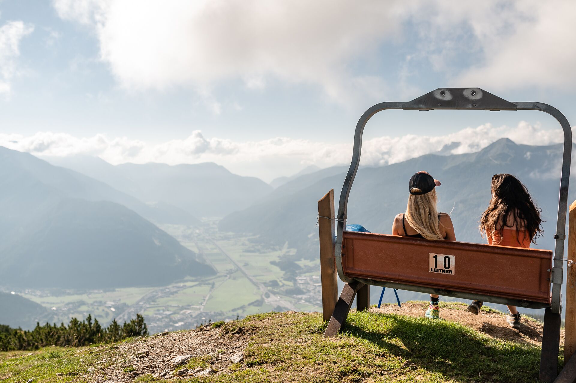 A bench with a sign reading "10 LEITNER" overlooking a mountainous landscape.
