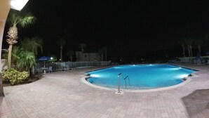 Uma área de piscina à noite, com deck pavimentado, espreguiçadeiras e palmeiras.