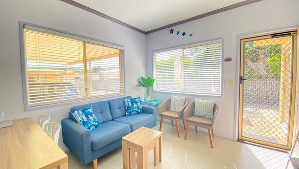 A room with a blue sofa, two wicker chairs, a small wooden table, and large windows with blinds. - San Marco Villa 3 - Burrum Heads - Beachfront - 2BR - Pool (BURRUM HEADS)