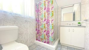 Une salle de bain comprenant des toilettes, un rideau de douche à motifs floraux, un lavabo avec un miroir au-dessus et un meuble sur lequel est posée une plante.