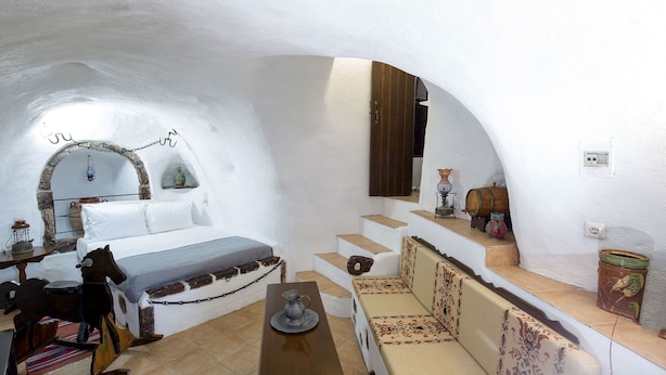 A cozy, white-walled room with a bed, a small table, and a sofa. There are steps leading to a higher level, and a wooden cabinet above the sofa. - Mylopetres (Santorini)
