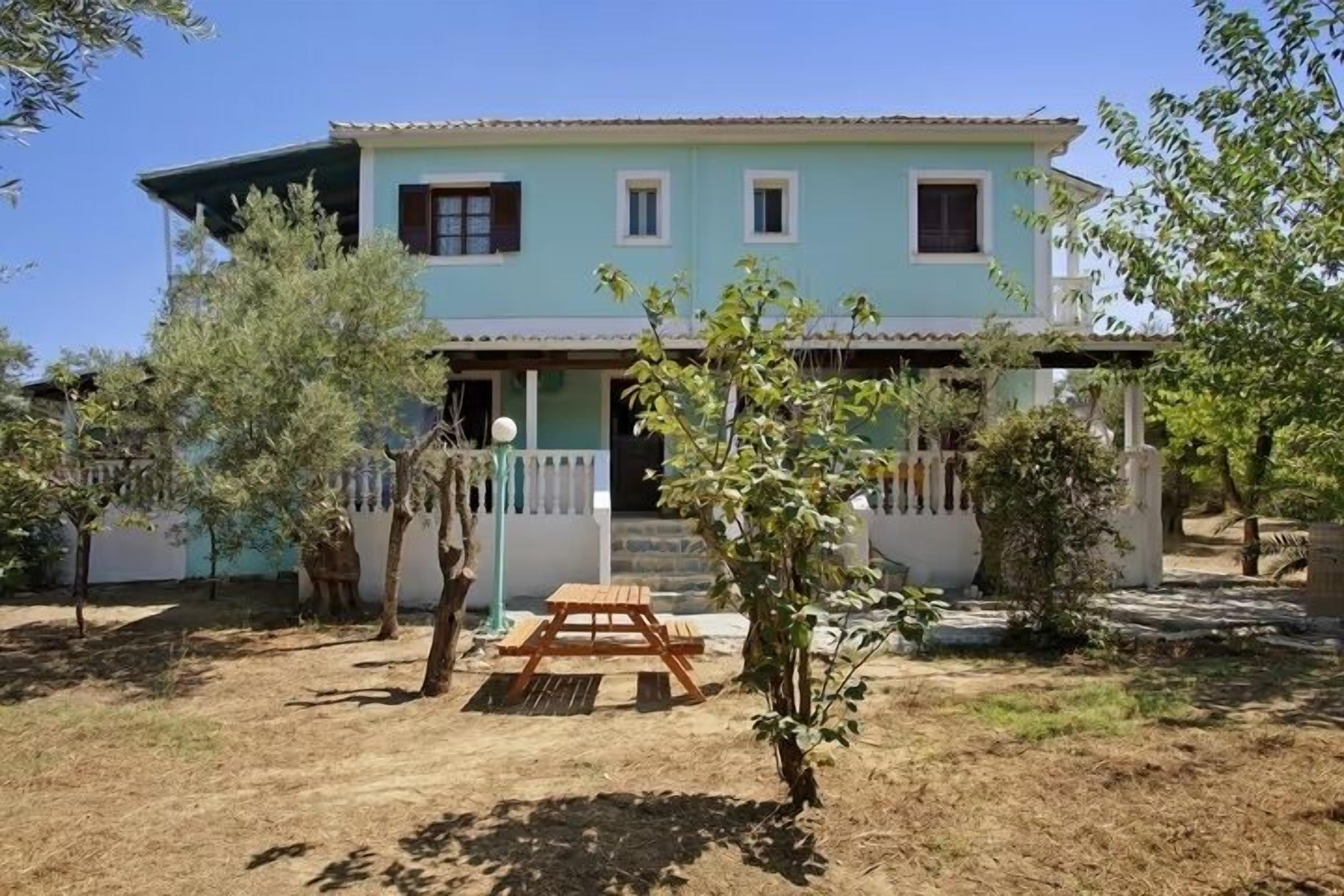 A two-story house with a light blue exterior, a wooden bench, and a small garden area with trees and shrubs.