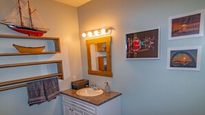A bathroom with a vanity, a ship model on a shelf, a mirror with lights, and framed pictures on the wall.