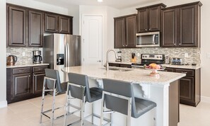 A modern kitchen with dark wood cabinets, stainless steel appliances, a granite countertop, and bar stools. - Vibrant Home with Private Pool in Encore (Kissimmee)