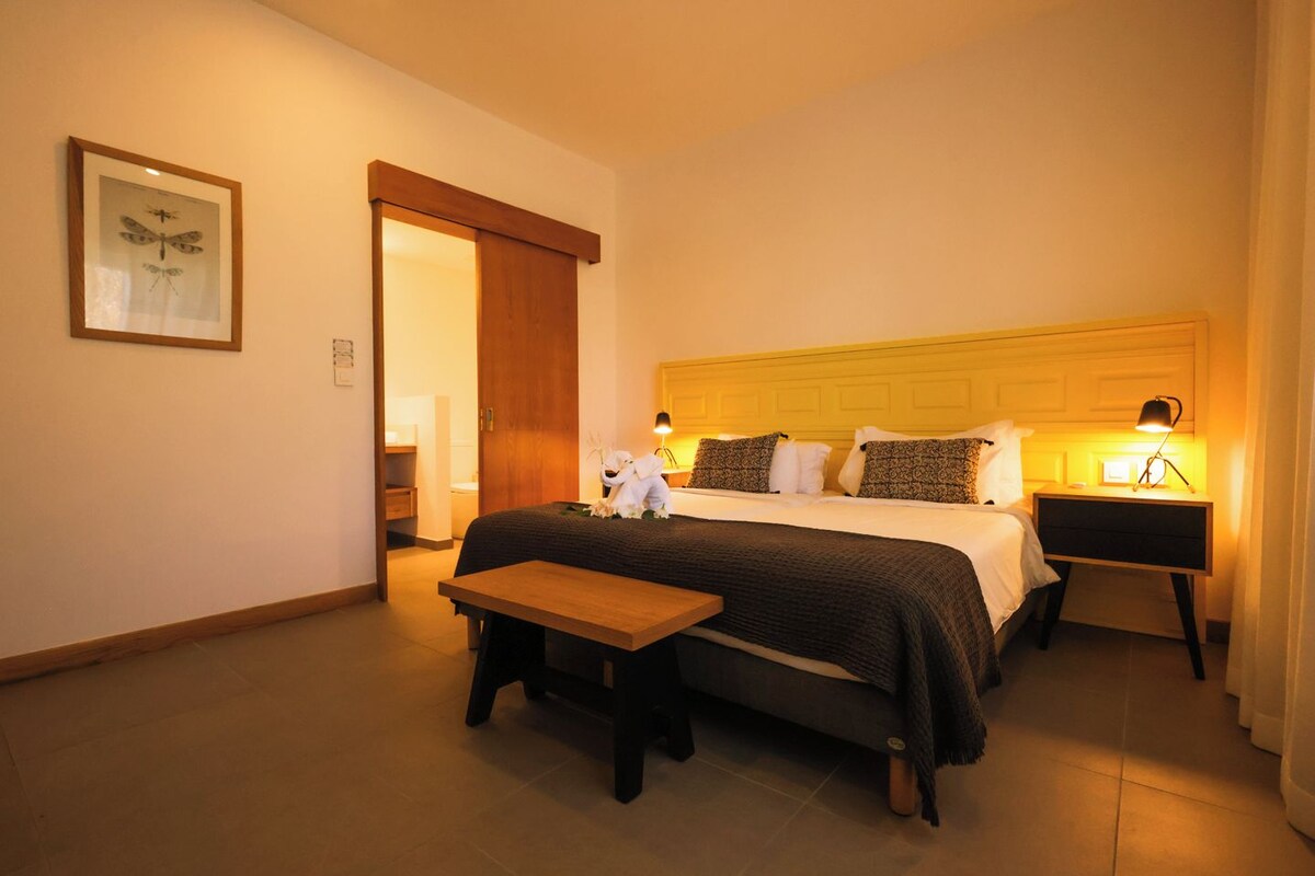 A hotel room with a bed, bedside tables, a bench, a framed picture on the wall, and a door leading to another room.