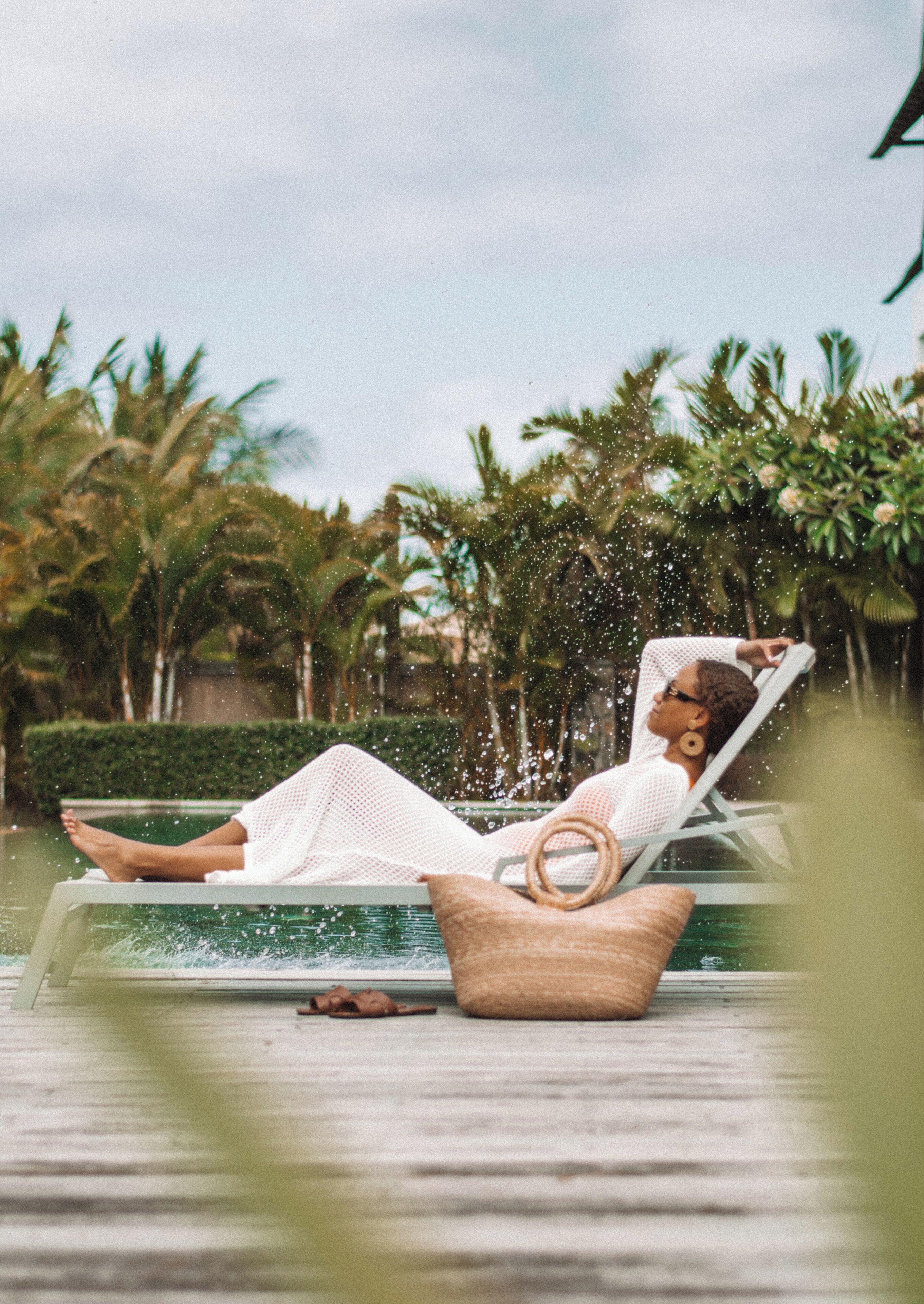 A person relaxing in a poolside chair with a wicker basket beside them, surrounded by lush greenery.