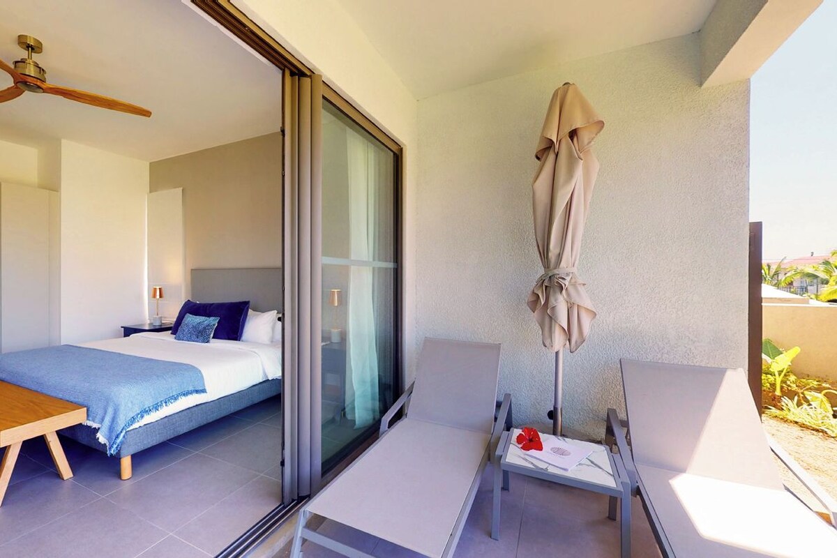 A modern bedroom with a bed, a wooden bench, a ceiling fan, and a sliding glass door leading to an outdoor area with a table and an umbrella.
