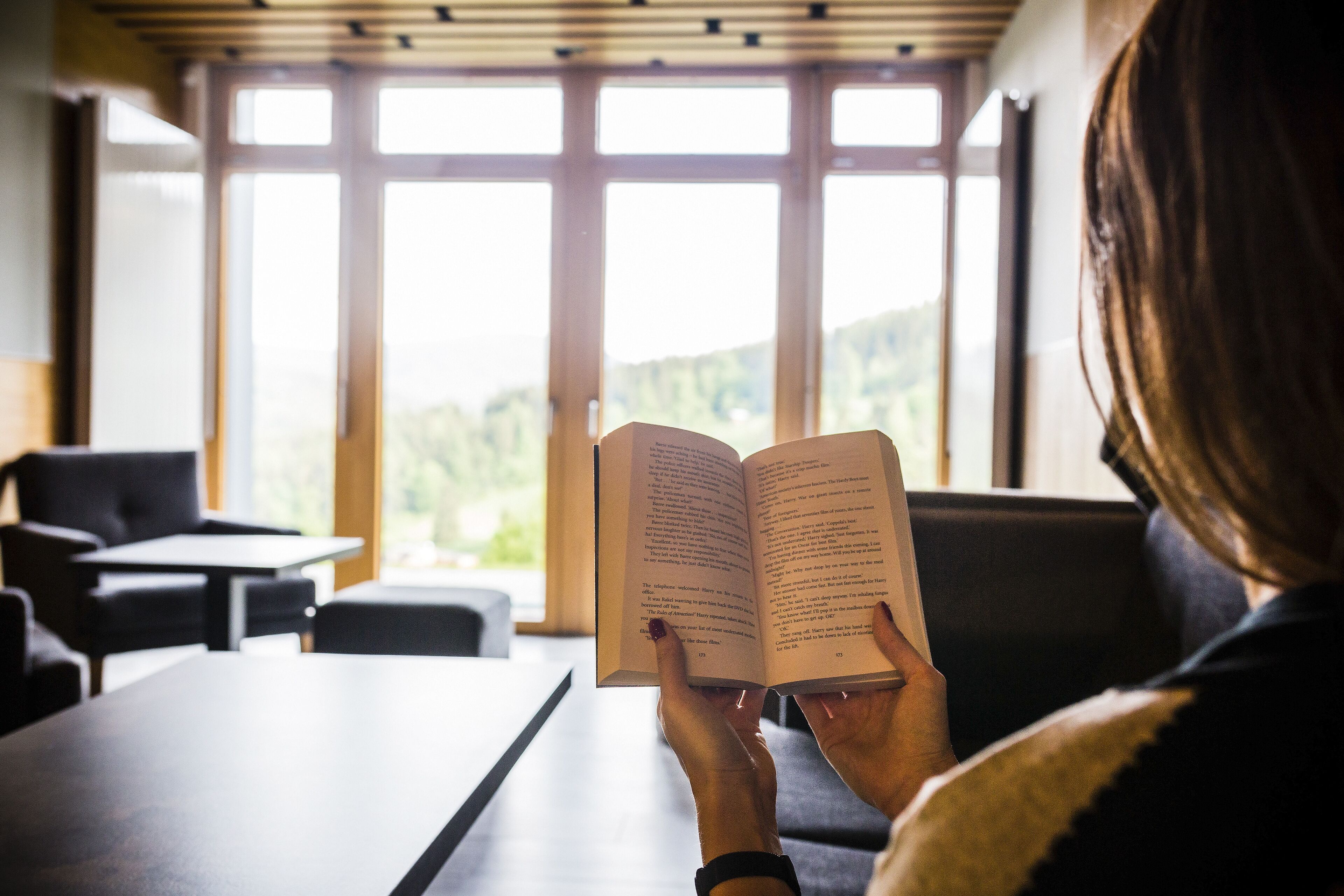 Una persona leyendo un libro en una habitación con ventanas amplias y vistas al exterior.