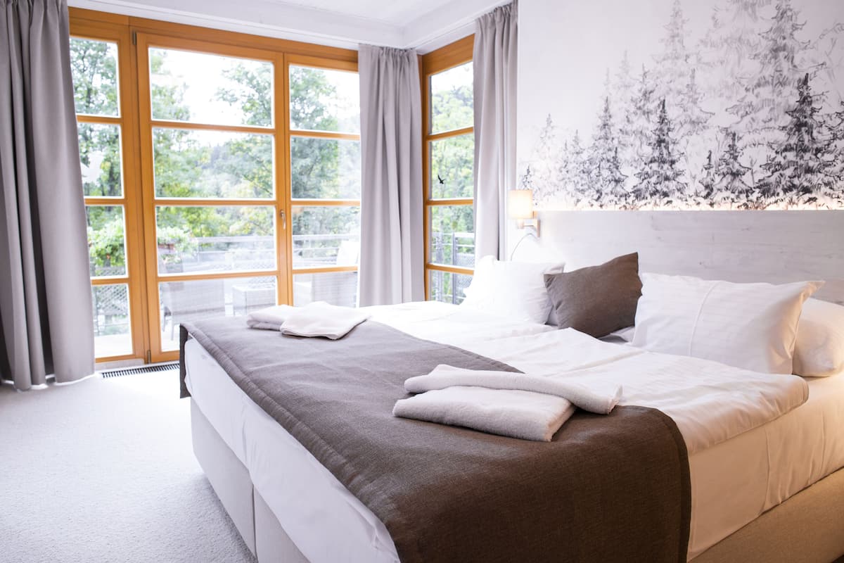 Een slaapkamer met een groot bed, grijze gordijnen en uitzicht op bomen door de ramen.