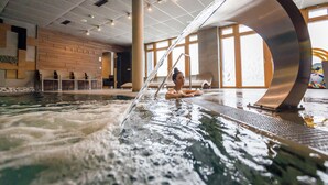 Une personne se détend dans un bain à remous, avec vue sur l’intérieur d’un bâtiment, notamment un mur en bois et une grande fenêtre.