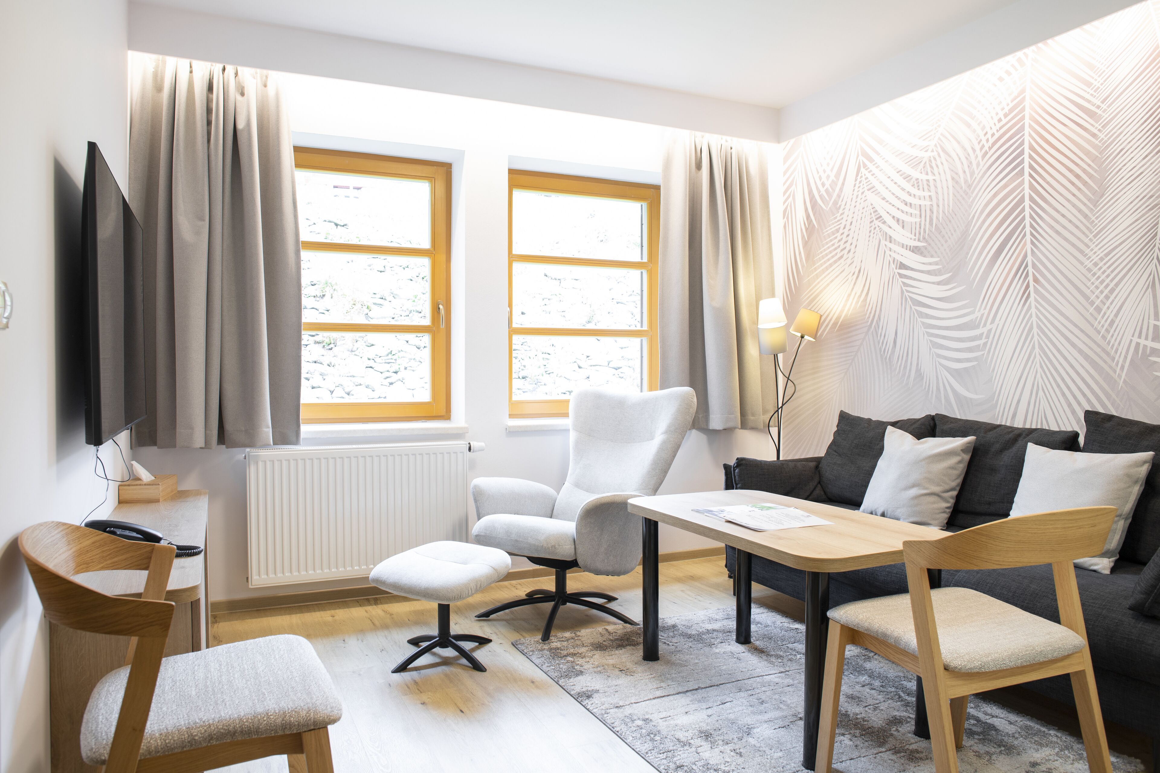 A modern living room with a sofa, chairs, a coffee table, and a TV. There are two windows with curtains, a radiator, and a wall with a leaf pattern.