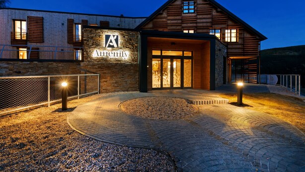 Un bâtiment moderne doté d’une façade en pierre et d’étages supérieurs en bois, avec une entrée imposante agrémentée de portes vitrées et une allée bordée de verdure.