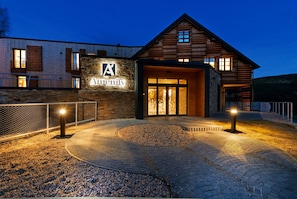 Un bâtiment moderne doté d’une façade en pierre et d’étages supérieurs en bois, avec une entrée imposante agrémentée de portes vitrées et une allée bordée de verdure.