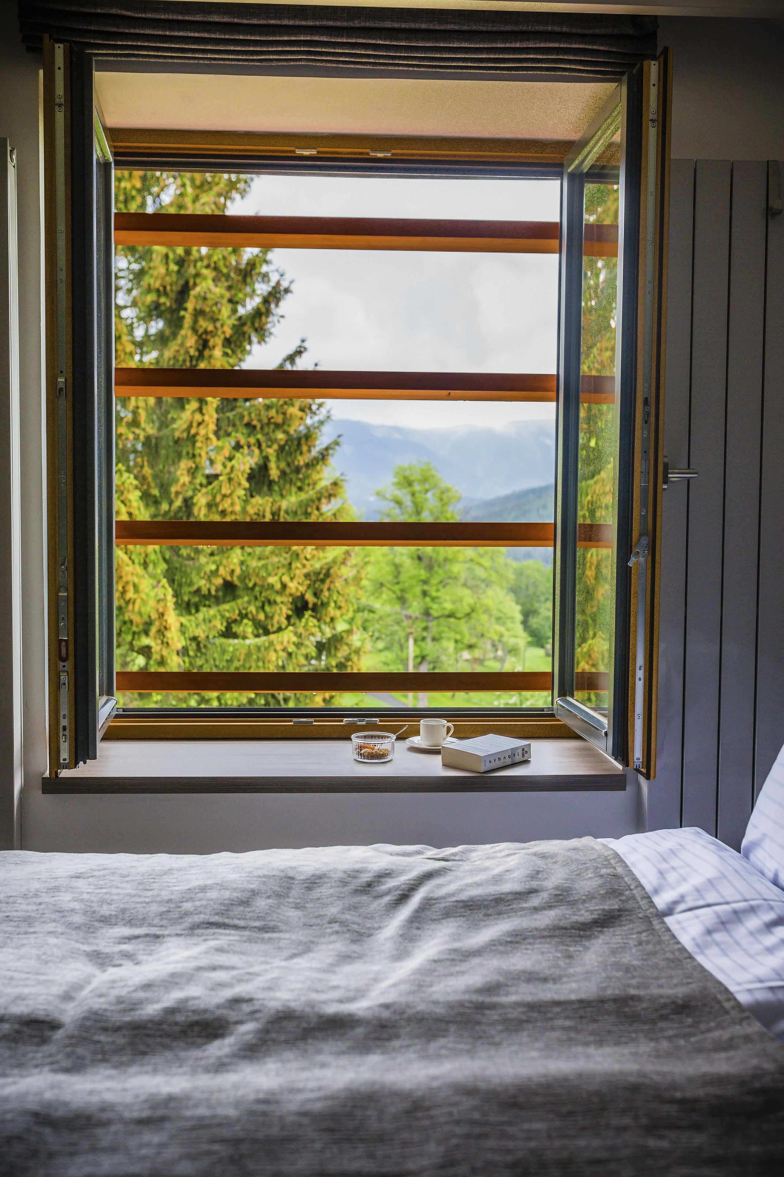 A bedroom with a large window, a bed, and a bedside table with a cup and a box.