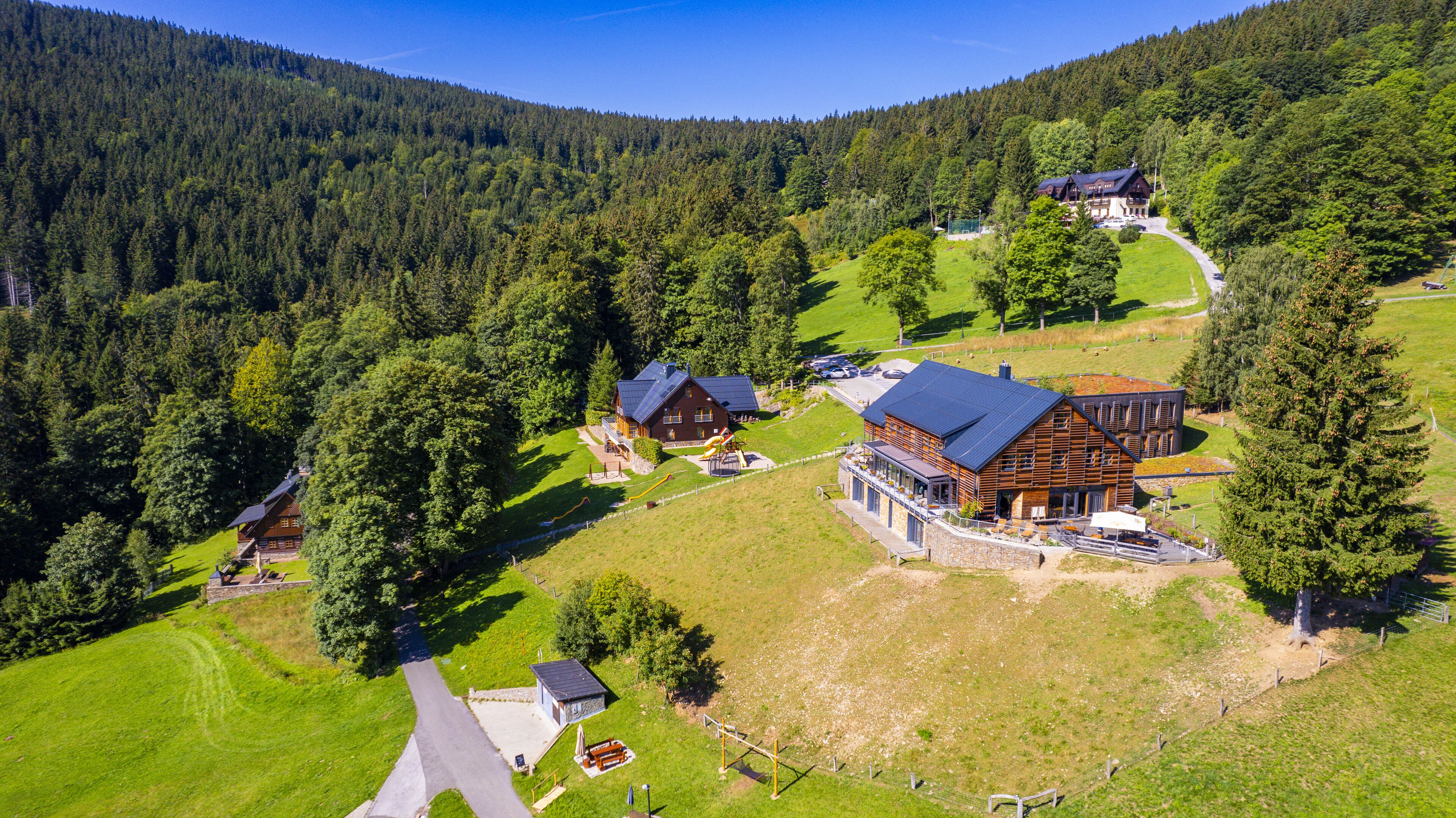 A large wooden house with a sloped roof, surrounded by a forested area and a small driveway.
