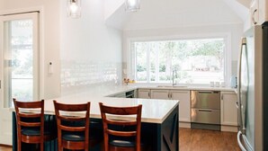 A kitchen with a large island, wooden chairs, stainless steel appliances, and a view of the backyard through large windows. - One Bedroom Cottage with a Shared Pool (Dural)