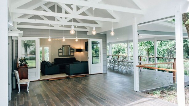 A spacious living area with a wooden floor, a dark-colored sofa set, and a large window offering a view of the outdoors. - One Bedroom Cottage with a Shared Pool (Dural)