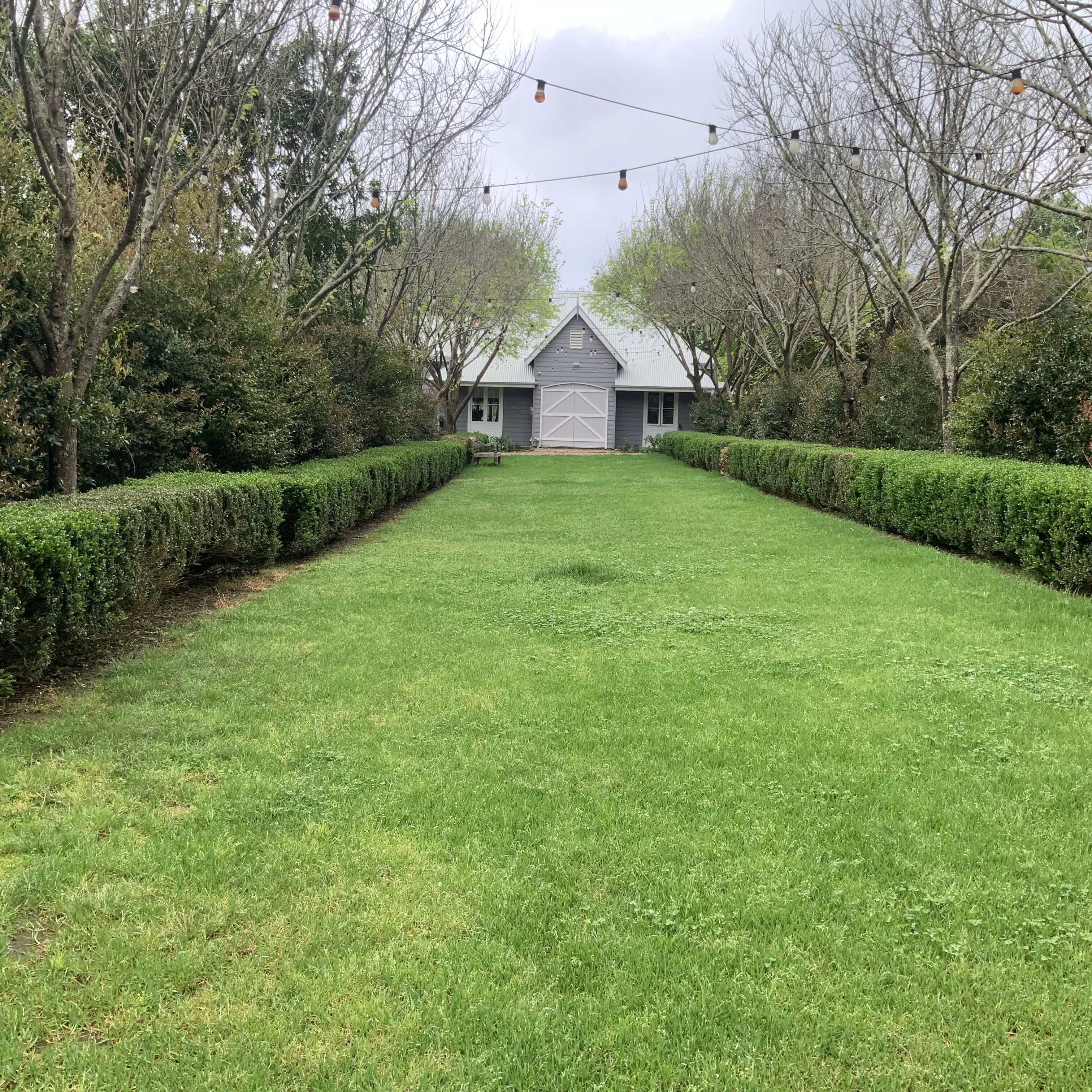 A well-maintained lawn with a symmetrical hedge on either side, leading to a building with a garage and a sloped roof.