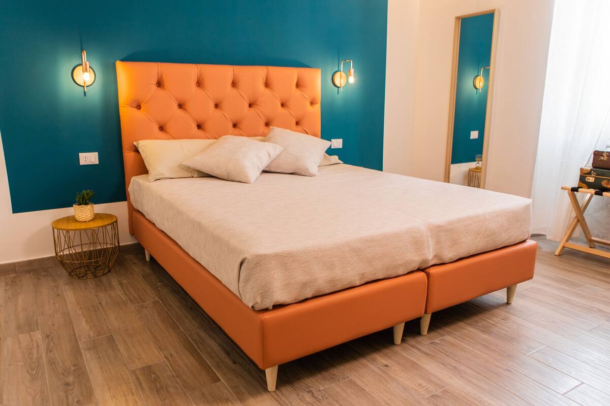 Uma cama com cabeceira laranja e roupa de cama branca, uma pequena mesa redonda com um cesto, e chão de madeira.